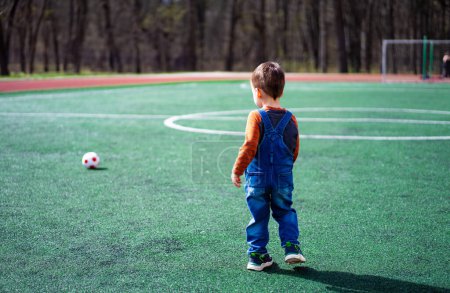 Téléchargez les photos : Un jeune garçon se tient sur un terrain vert avec un ballon de football devant lui. Il porte une chemise rouge et une salopette bleue - en image libre de droit