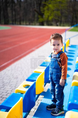 Téléchargez les photos : Un jeune garçon se tient debout sur un gradin avec un siège bleu et jaune derrière lui. Il porte une chemise bleue et une chemise orange - en image libre de droit