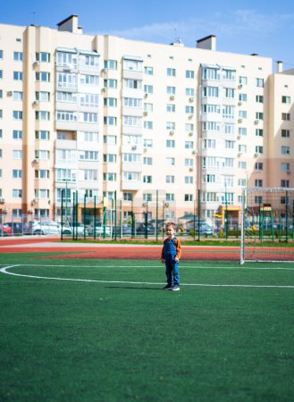 Téléchargez les photos : A young boy stands on a green field in front of a large apartment building. The boy is wearing a blue shirt and jeans - en image libre de droit
