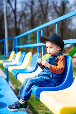 Téléchargez les photos : A young boy is sitting on a blue and yellow bench, eating a drink from a plastic cup - en image libre de droit