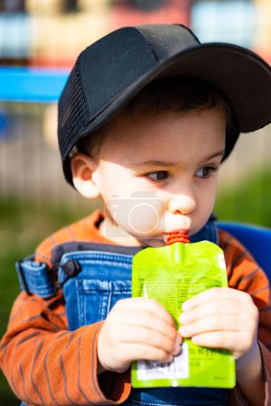 Téléchargez les photos : A young boy is sitting on a blue and yellow bench, eating a drink from a plastic cup - en image libre de droit