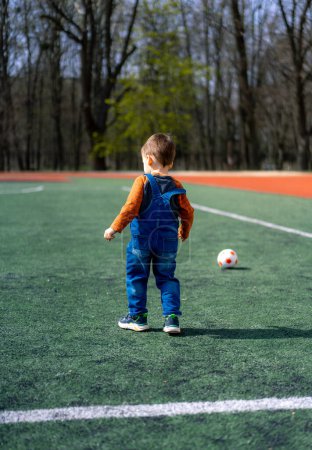 Téléchargez les photos : A young boy stands on a soccer field with a soccer ball and a second ball. The boy is wearing a blue overalls and an orange shirt - en image libre de droit