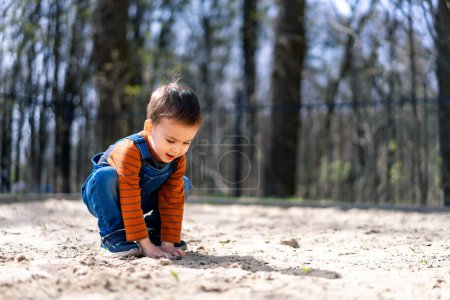 A young boy is playing in the sand, wearing a blue shirt and orange pants. He is smiling and he is enjoying himself