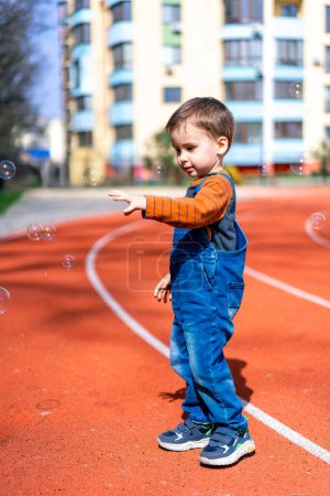 Téléchargez les photos : Un jeune garçon est debout sur une piste avec des bulles dans l'air. Il pointe vers les bulles, et la scène a une ludique - en image libre de droit