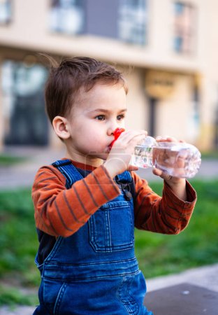 Téléchargez les photos : Un jeune garçon boit l'eau d'une bouteille. Il porte une salopette bleue et une chemise rayée - en image libre de droit