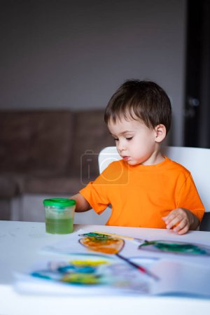 Téléchargez les photos : Un jeune garçon est assis à une table avec un récipient vert de pâte à jouer devant lui. Il dessine sur un morceau de papier avec un marqueur vert - en image libre de droit
