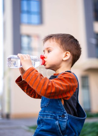 Téléchargez les photos : A young boy is drinking water from a bottle. The bottle has a red cap. The boy is wearing an orange shirt and blue overalls - en image libre de droit