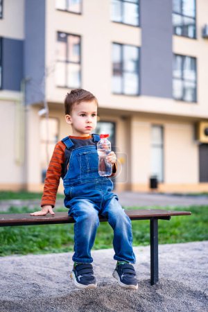 Téléchargez les photos : Un jeune garçon s'assoit sur un banc d'eau potable. Le garçon porte une salopette bleue et une chemise rayée. - en image libre de droit