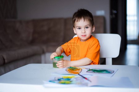 Téléchargez les photos : Un jeune garçon en chemise orange s'assoit à une table avec un pot de peinture verte. - en image libre de droit