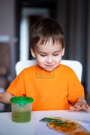 Téléchargez les photos : Un jeune garçon en chemise orange s'assoit à une table avec une coupe verte devant lui - en image libre de droit
