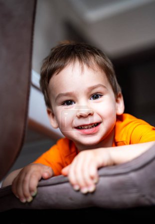 Téléchargez les photos : Un jeune garçon en chemise orange sourit et regarde la caméra. Il est assis sur un canapé et il s'amuse - en image libre de droit