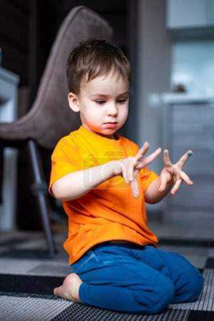 Téléchargez les photos : A young boy in an orange shirt is playing with his hands. He is sitting on the floor and he is focused on his activity - en image libre de droit