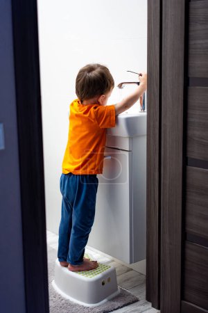 Téléchargez les photos : A young boy is standing on a stool in front of a sink. He is washing his hands - en image libre de droit