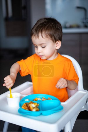 Téléchargez les photos : Un jeune garçon est assis sur une chaise haute à manger de la nourriture. Il porte une chemise orange et a une cuillère à la main - en image libre de droit
