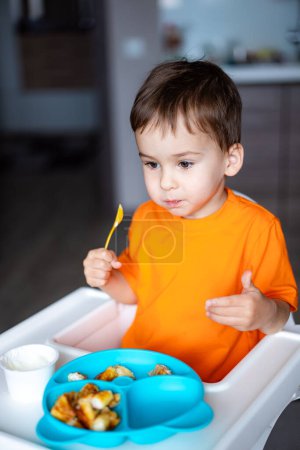 Téléchargez les photos : Un jeune garçon mange de la nourriture dans une assiette bleue. Il porte une chemise orange et a une cuillère à la main - en image libre de droit
