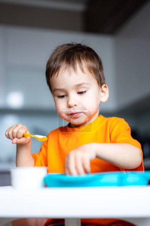 Téléchargez les photos : Un jeune garçon mange un bol de nourriture avec une cuillère. Il porte une chemise orange. - en image libre de droit
