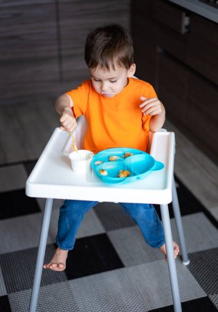 Téléchargez les photos : A young boy is sitting in a high chair eating food. He is wearing an orange shirt and has a spoon in his mouth - en image libre de droit