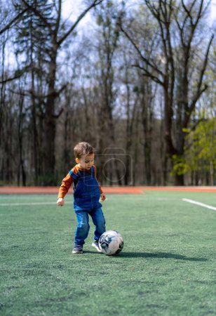 Photo for A young boy in blue overalls kicks a soccer ball on a green field. The boy is wearing a red shirt and has a smile on his face - Royalty Free Image