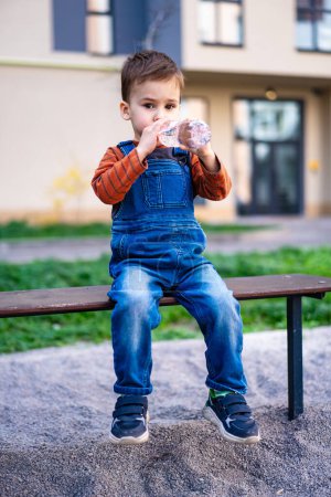 Téléchargez les photos : Un jeune garçon s'assoit sur un banc d'eau potable. Le garçon porte des salopettes bleues et une chemise rayée - en image libre de droit