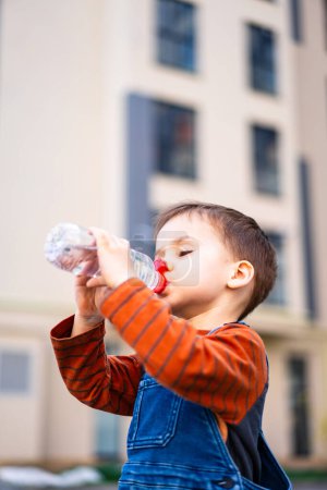 Téléchargez les photos : Un jeune garçon boit l'eau d'une bouteille en plastique. La bouteille est rouge et blanche. Le garçon porte une chemise orange et une salopette bleue - en image libre de droit