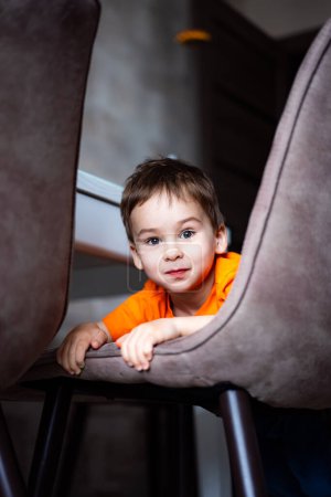 Téléchargez les photos : Un jeune garçon en chemise orange est assis sur une chaise. Il regarde la caméra avec un sourire sur son visage - en image libre de droit