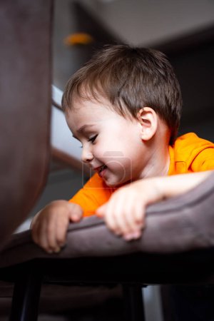 Téléchargez les photos : Un jeune garçon en chemise orange est assis sur une chaise. Il sourit et regarde quelque chose. - en image libre de droit