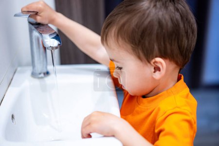 Téléchargez les photos : Un jeune garçon joue avec l'eau dans un évier. Il porte une chemise orange. L'évier est blanc et a un robinet en argent - en image libre de droit