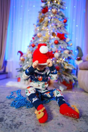 Téléchargez les photos : Un enfant est assis devant un sapin de Noël portant un chapeau rouge - en image libre de droit