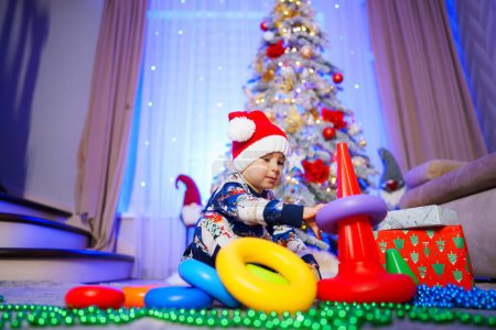 Téléchargez les photos : Un jeune enfant joue avec un ensemble d'anneaux en plastique devant un sapin de Noël. - en image libre de droit