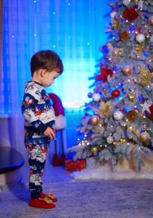 Téléchargez les photos : Un jeune garçon en pyjama se tient devant un sapin de Noël. La chambre est décorée avec des lumières et des cadeaux, créant une atmosphère festive - en image libre de droit