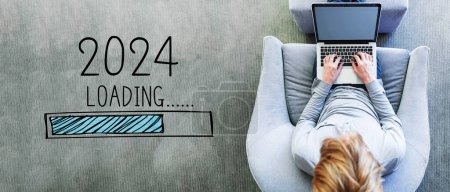 Téléchargez les photos : Chargement de la nouvelle année 2024 avec l'homme en utilisant un ordinateur portable dans une chaise grise moderne - en image libre de droit