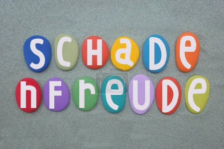 Schadenfreude, the experience of pleasure, joy, or self-satisfaction that comes from learning of or witnessing the troubles, failures, pain, or humiliation of another, text composed with hand painted multi colored stone letters over green sand