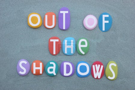 Out of shadows, creative text composed with hand painted multi colored stone letters over green sand