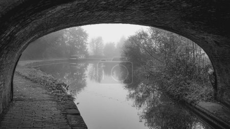 En regardant sous un pont de briques au-dessus du canal, l'arche encadre la vue en bas du canal où un bateau étroit peut être vu dans la brume. L'image est en noir et blanc