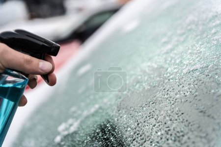 Foto de El hombre usa una botella de descongelador para descongelar el parabrisas cubierto de hielo de su coche. - Imagen libre de derechos