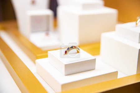 Foto de Anillo de oro con piedras preciosas de colores presentado en un cojín blanco con un fondo borroso de exhibición de joyas - Imagen libre de derechos