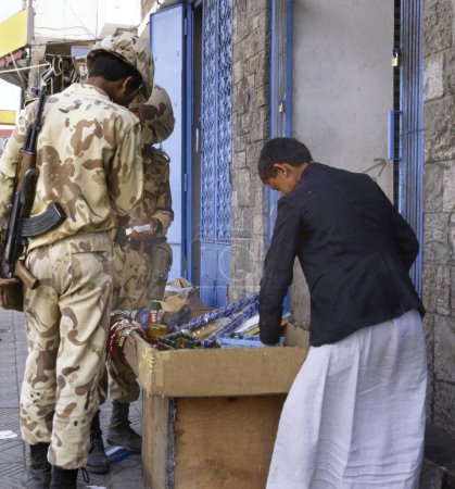 Téléchargez les photos : SANA'A, YEMEN- 15 AVRIL 2009 : Jeune vendeur de cigarettes et d'autres matériaux dans une rue vendant du tabac à des soldats avec des kalachkinovs - en image libre de droit
