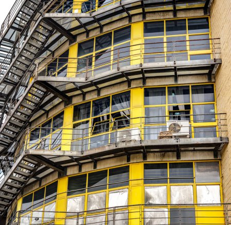 Foto de Un antiguo edificio industrial con prominentes escaleras de emergencia y grandes ventanas amarillas, mostrando su diseño arquitectónico histórico. - Imagen libre de derechos