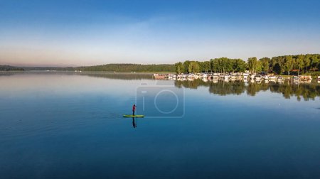 Photo for Woman paddling on SUP board on beautiful lake aerial drone view with reflections from above. Standing up paddle boarding adventure in early morning sunrise. Germany lake district Mecklenburg. - Royalty Free Image