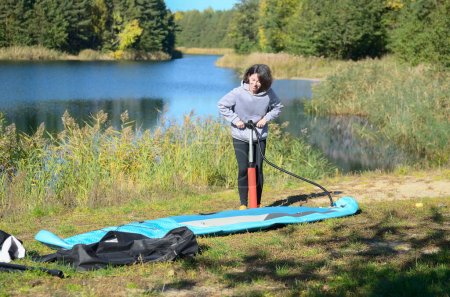 Photo for Active woman inflates SUP board with manual pump, beautiful lake and nature on background, stand up paddling water adventure outdoors, sport and healthy lifestyle concept - Royalty Free Image