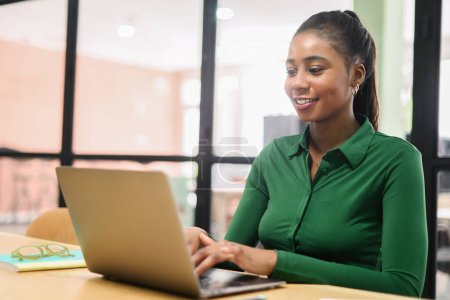 Portrait of young African American woman sitting at the desk with a laptop, typing an email, working, chatting, browsing online with a pleasant smile Poster 619025388