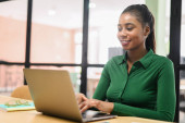Portrait of young African American woman sitting at the desk with a laptop, typing an email, working, chatting, browsing online with a pleasant smile Poster #619025388