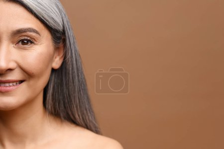 Téléchargez les photos : Gros plan portrait de la moitié du visage de gaie femme asiatique d'âge moyen des années 50 aux cheveux longs gris argenté, jolie dame regarde la caméra et rit. Modèle mature aux épaules nues isolé sur brun - en image libre de droit