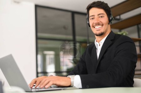 Téléchargez les photos : Friendly and smiling hispanic man wearing wireless headset looks at the camera sitting at the desk in front of laptop in modern office, male employee using hands free device for online connection - en image libre de droit