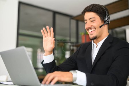 Handsome young hispanic businessman in smart formal business wear using headset and laptop for video communication, customer service representative greeting client, waving, saying hello
