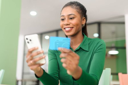 Foto de Excited African-American woman shopping online, holding banking credit card and smartphone, inputs data and making purchase online, multiracial female using e-banking app - Imagen libre de derechos