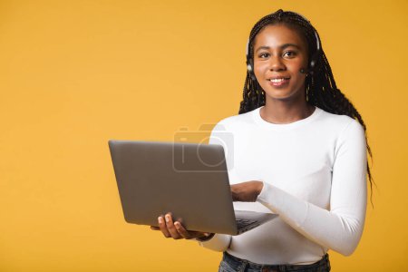 Foto de Sonriente empleada del centro de llamadas femenino usando un auricular y un portátil para la comunicación en línea con los clientes, mujer hablando en el micrófono y escribiendo en el teclado, cheques y marcas - Imagen libre de derechos
