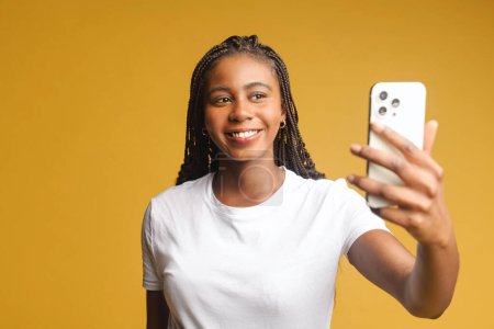 Téléchargez les photos : Happy carefree African-American young woman taking selfie on the smartphone, recording video message, streaming in social medias, isolated on yellow background - en image libre de droit