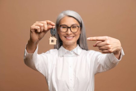Téléchargez les photos : Smiling woman pointing at the keys from new property, happy buyer of own estate. Happy overjoyed senior woman holding keys with keychain in form of little house isolated on brown. Lady relocated - en image libre de droit