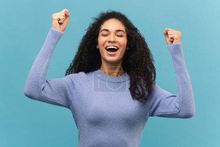 Téléchargez les photos : Portrait of excited overjoyed fashionably dressed woman standing with raised hands and shouting yeah, Im winner, rejoicing victory, success. Studio shot isolated on blue background - en image libre de droit